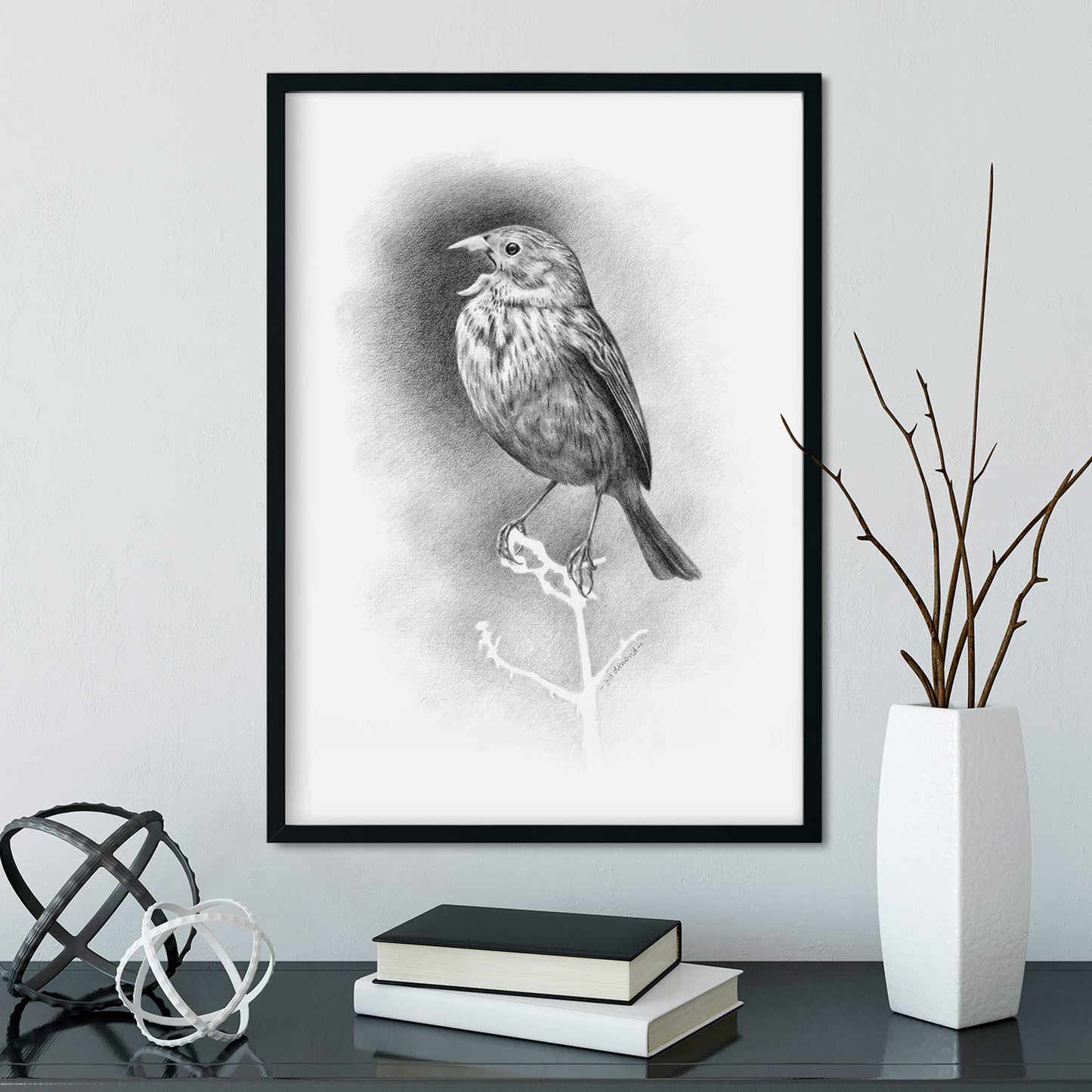 Black frame on a wall containing a black and white drawing of a small bird singing. Desk below with vase and books