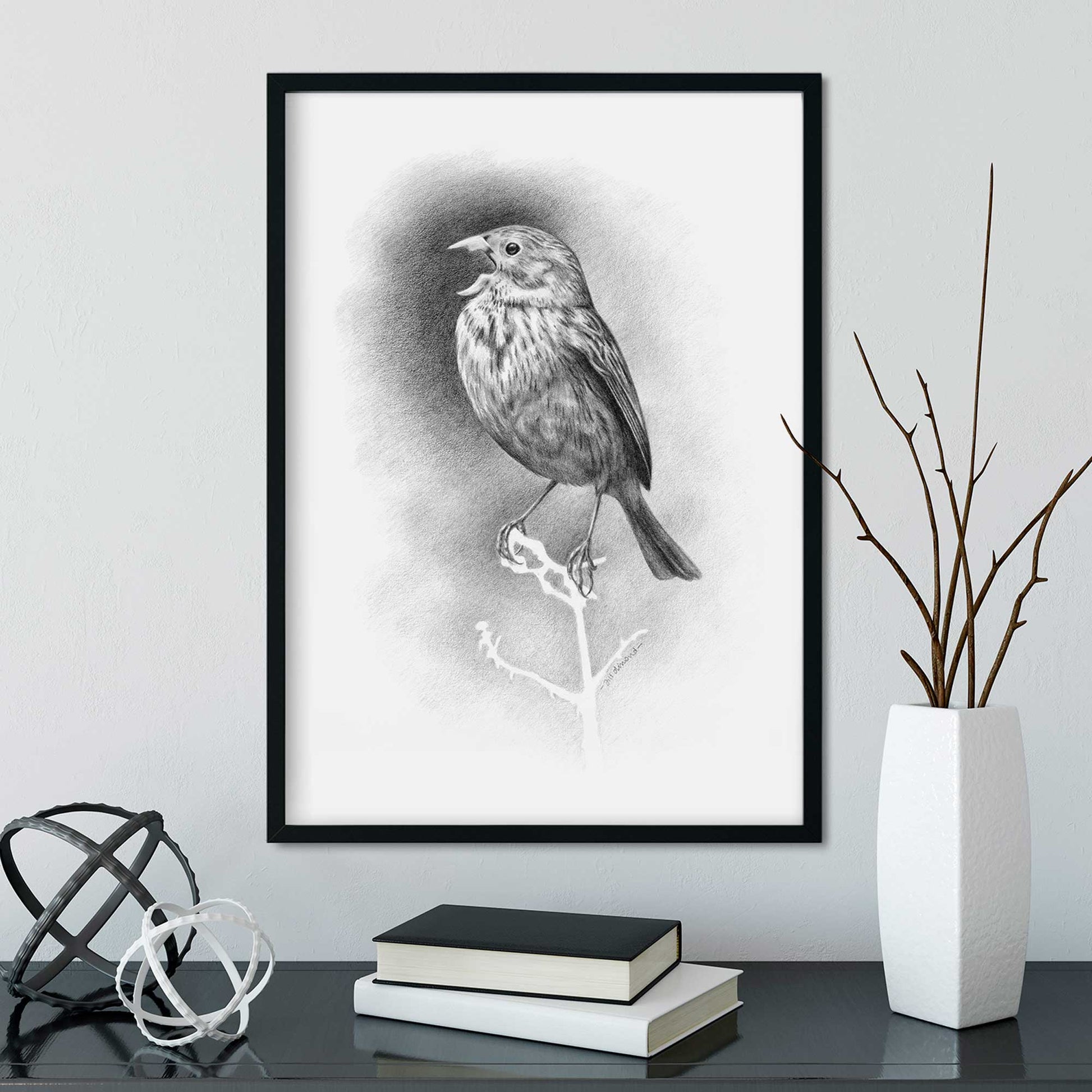 Black frame on a wall containing a black and white drawing of a small bird singing. Desk below with vase and books