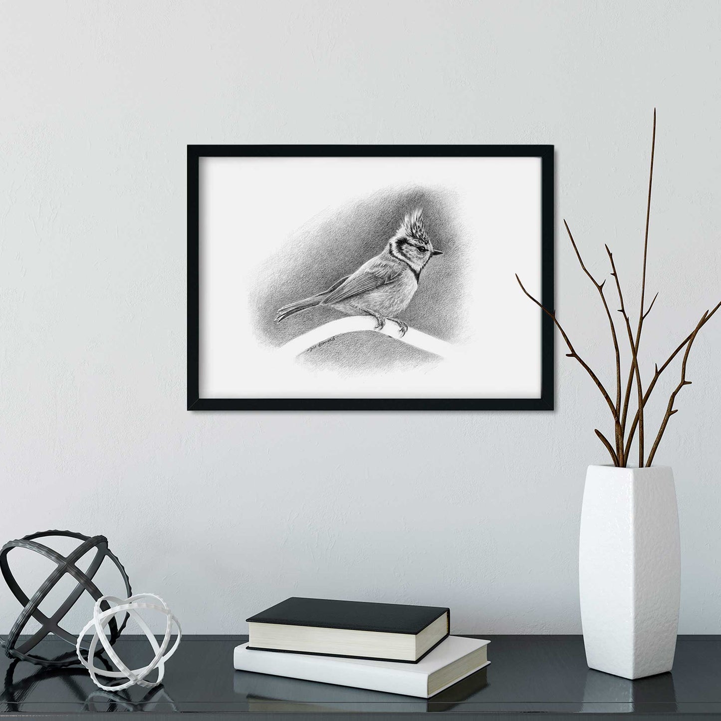 Black frame on wall containing black and white drawing of a crested tit bird. desk below with vase and books