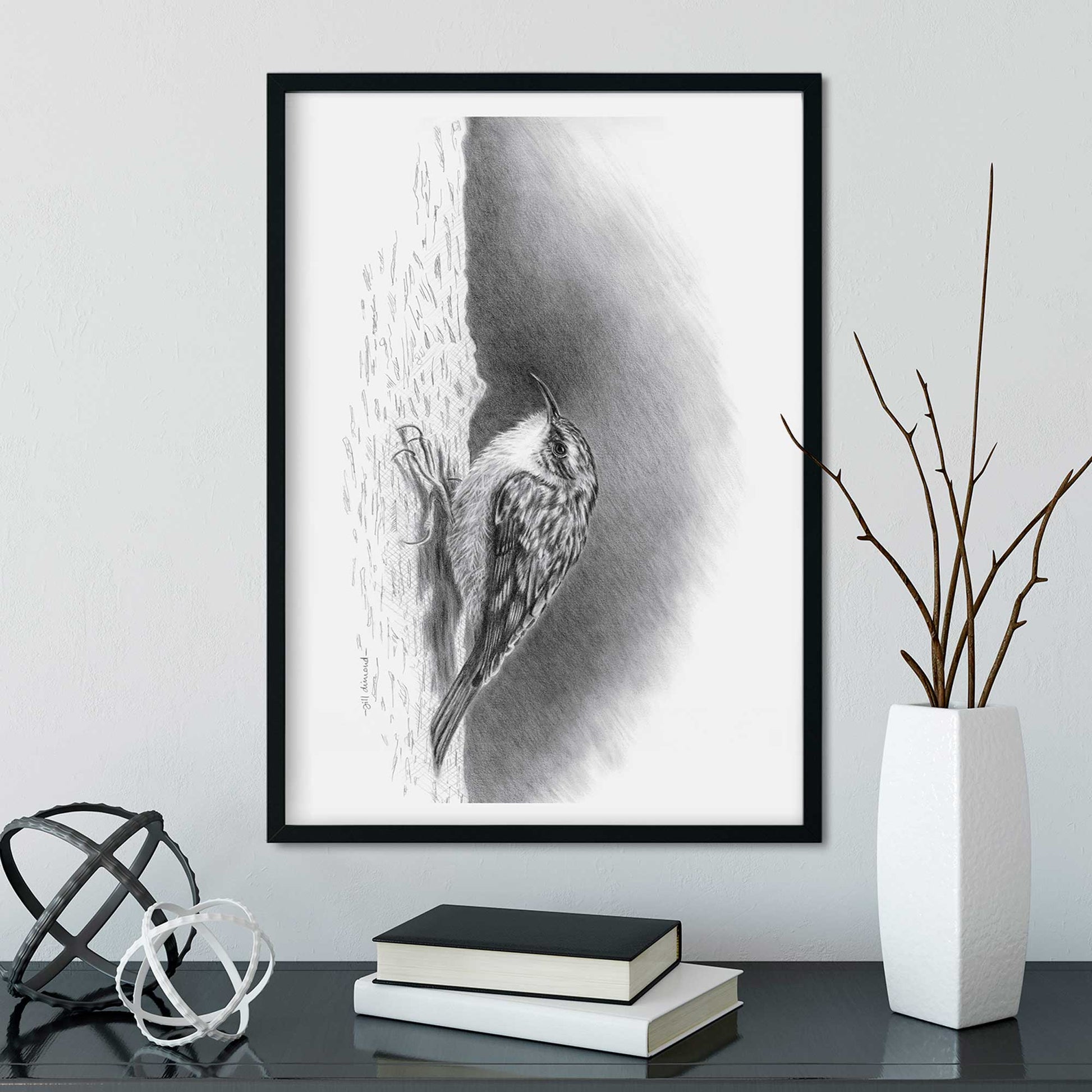 Black frame on wall containing drawing of a small treecreeper bird.  Desk below with vase and books