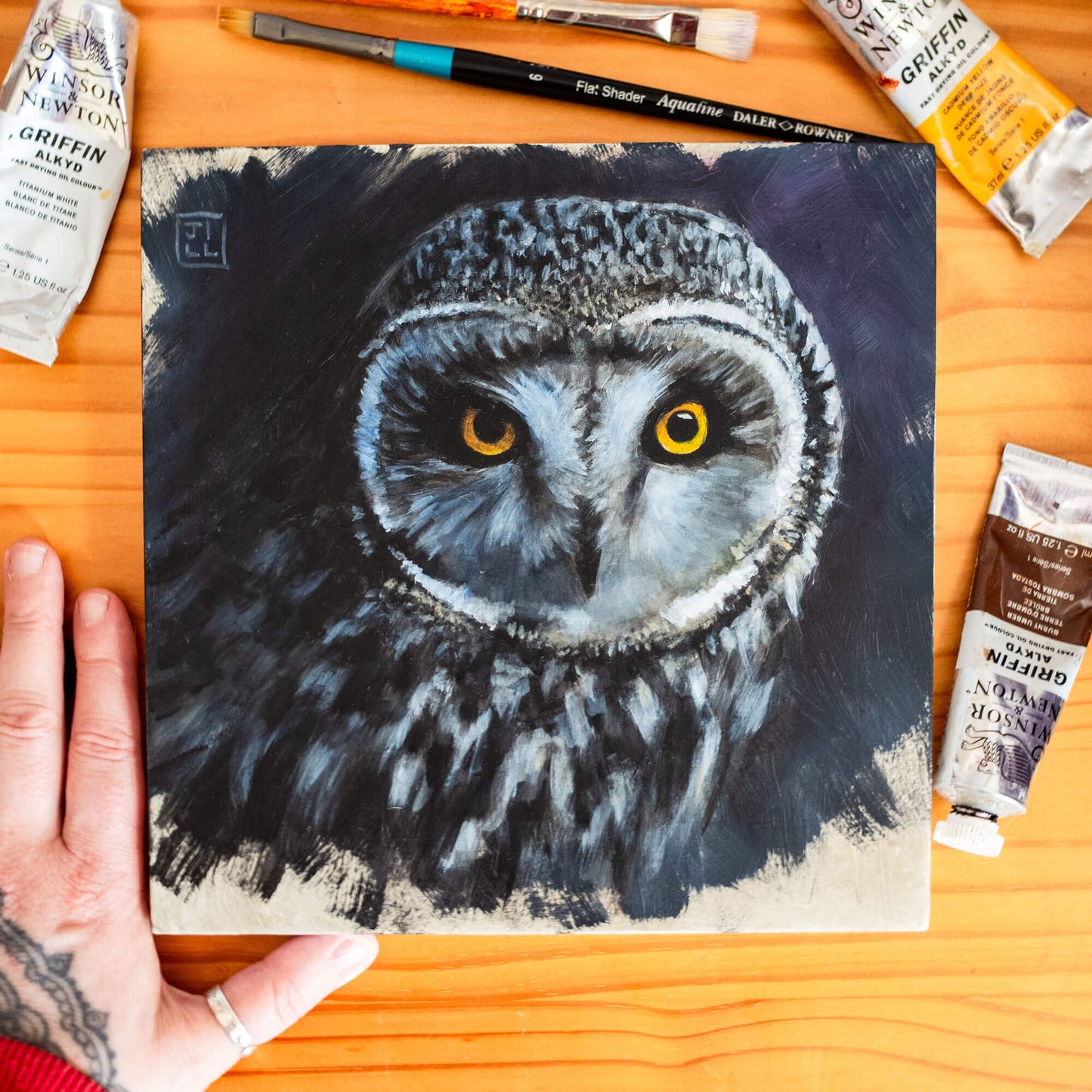 Hand holding a painting of a short-eared owl on a wooden surface with art supplies.
