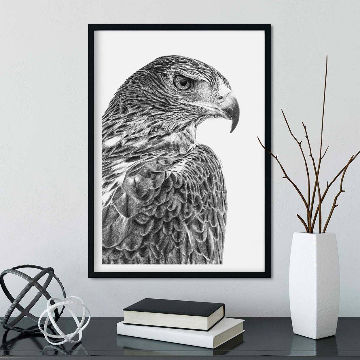 Black frame on wall containing black and white drawing of a golden eagle.  Desk below with books and vase on it.