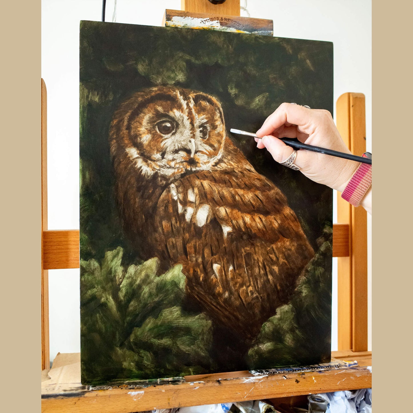 Tawny owl original oil painting work in progress by bird artist Jill Dimond, with the painting on an easel and the artist's hand holding a brush